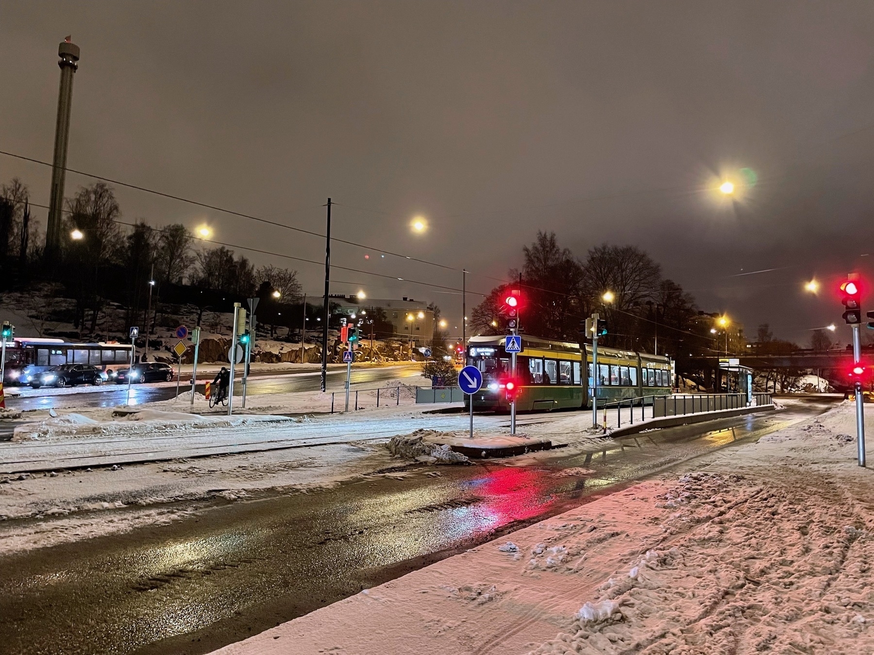 An intersection with several traffic lights, a tram waiting at a red light, some traffic in the background and snow on the sidewalks.