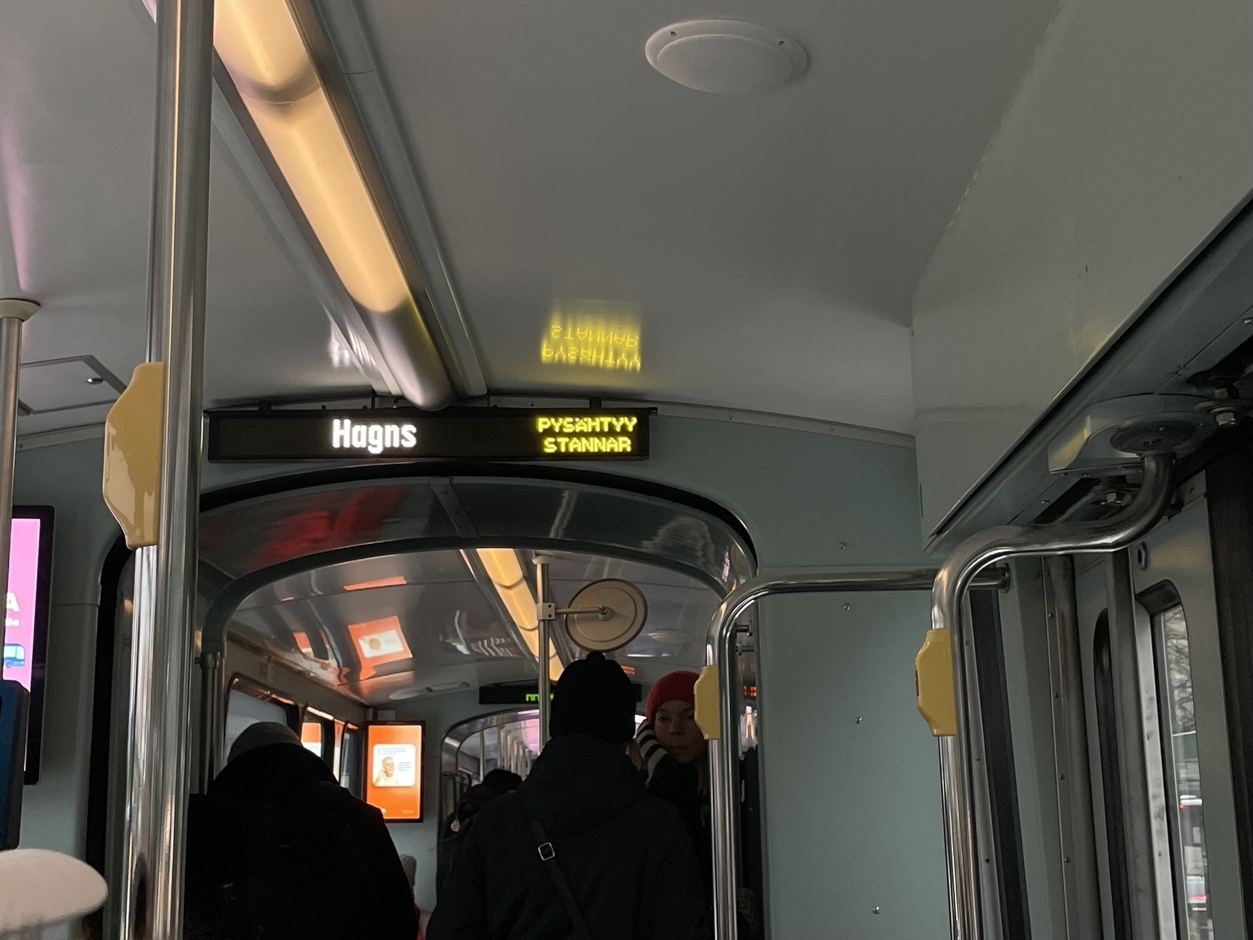 Tram interior with sign saying “Hagns” as well as ”pysähtyy” and ”stannar”. There are some people on the tram. 