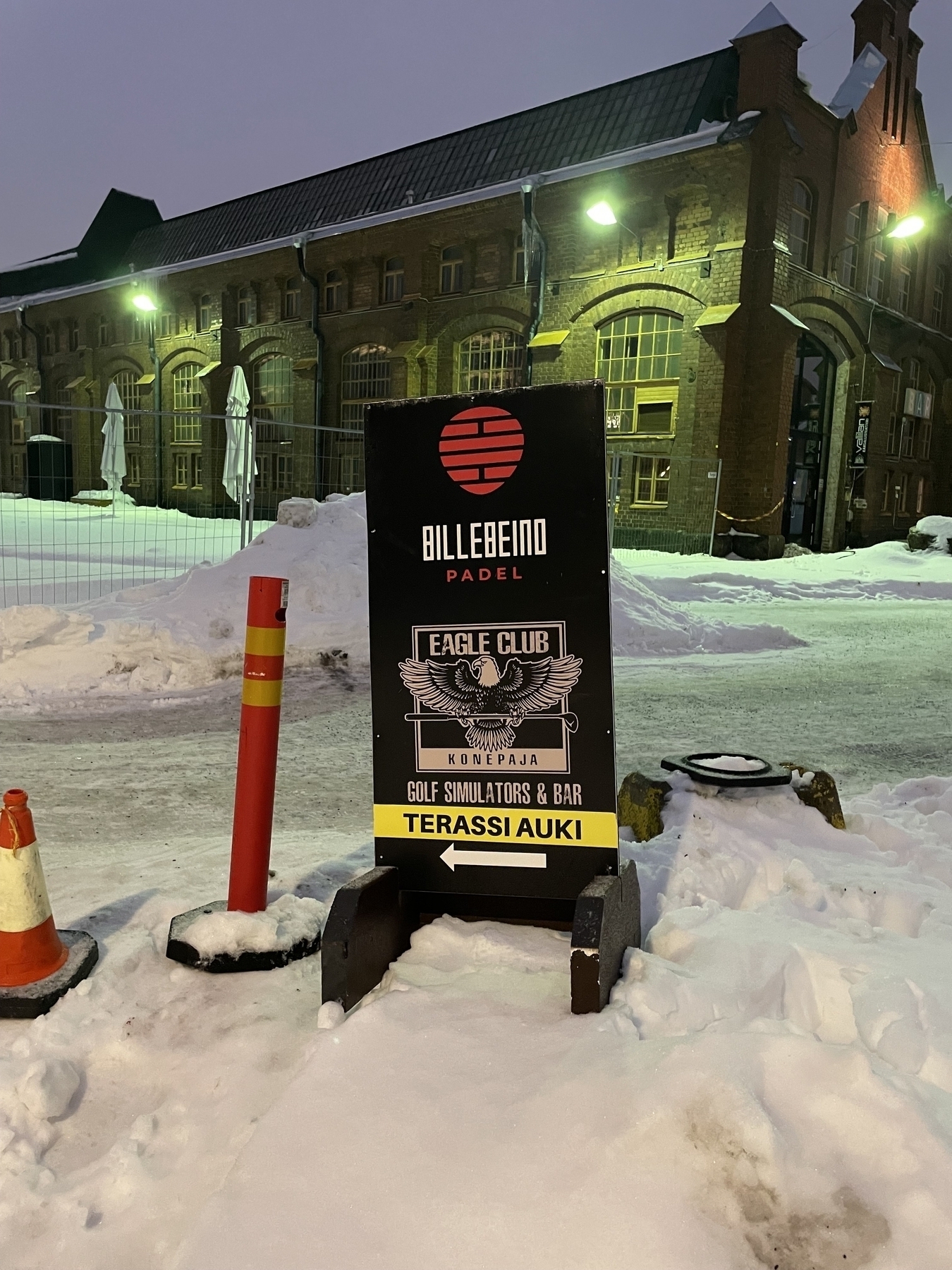 A sign saying “EAGLE CLUB Golf Simulators & pub. TERASSI AUKI”. The sign is standing in a pile of snow. 