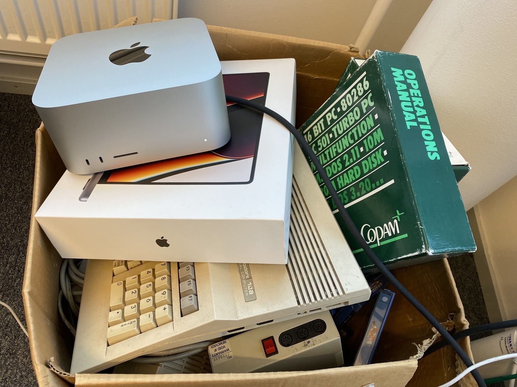 An overflowing cardboard box - on the top of the pile is a Mac Studio (powered on), sitting on top of a MacBook box, sitting on top of a Commodore 128. Next to them is a computer manual in a thick green binder labelled “OPERATIONS MANUAL” and “16 BIT PIC • 80286 • TURBO PC • … DOS 2.11 • … 10 M … HARD DISK • DOS 3.20…”